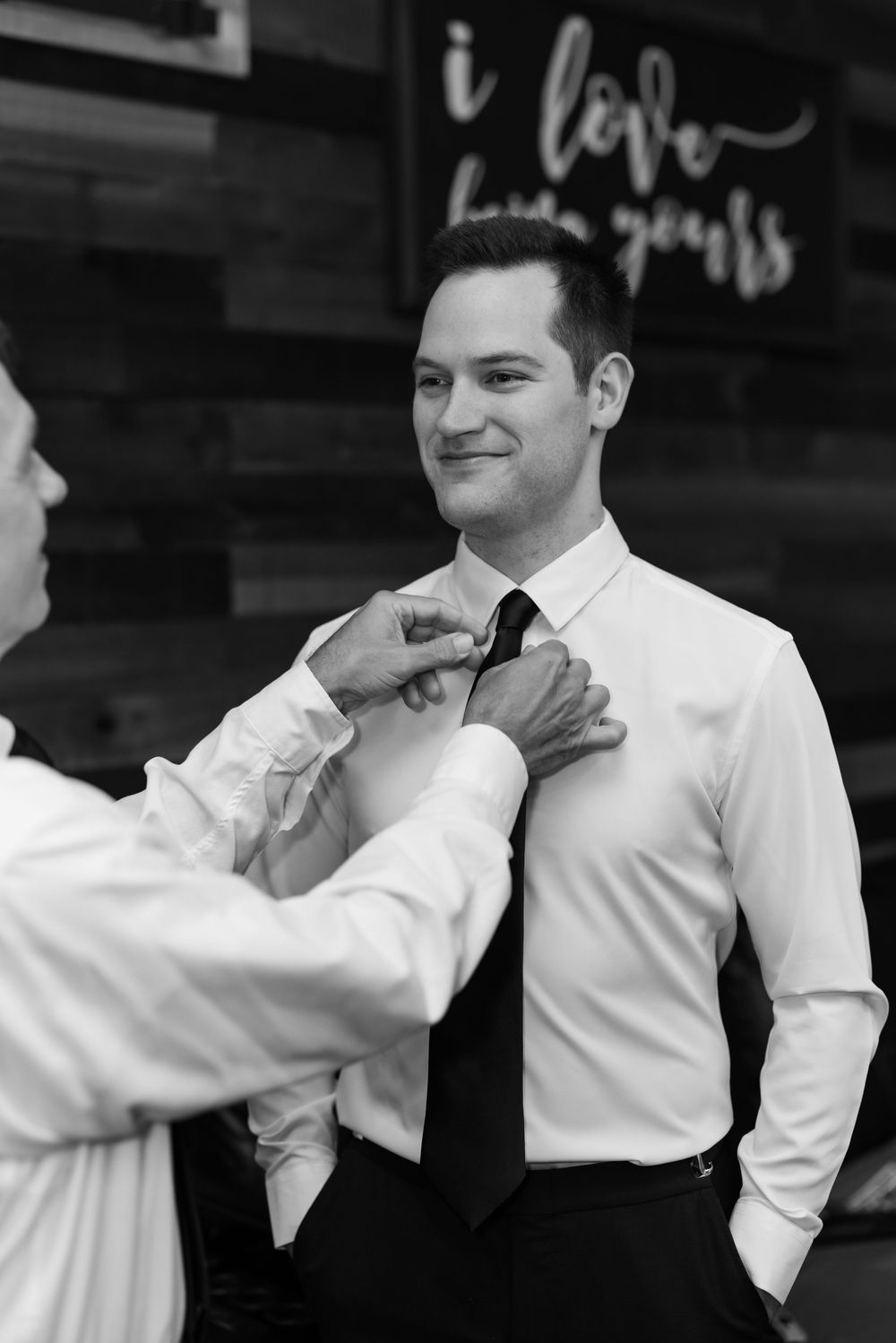 Black and white photo of a groom smiling as someone adjusts his tie before the wedding.