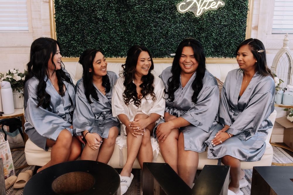 Bride in white robe laughing with four bridesmaids in silver robes seated on a couch during wedding preparations.