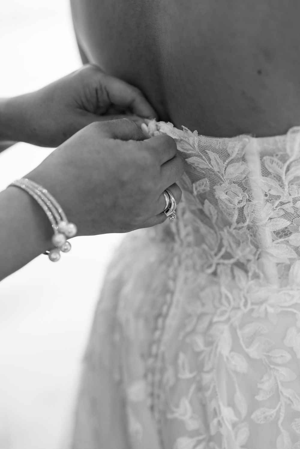 Hands fastening lace wedding dress buttons, with pearl bracelet and ring visible in black and white photo.