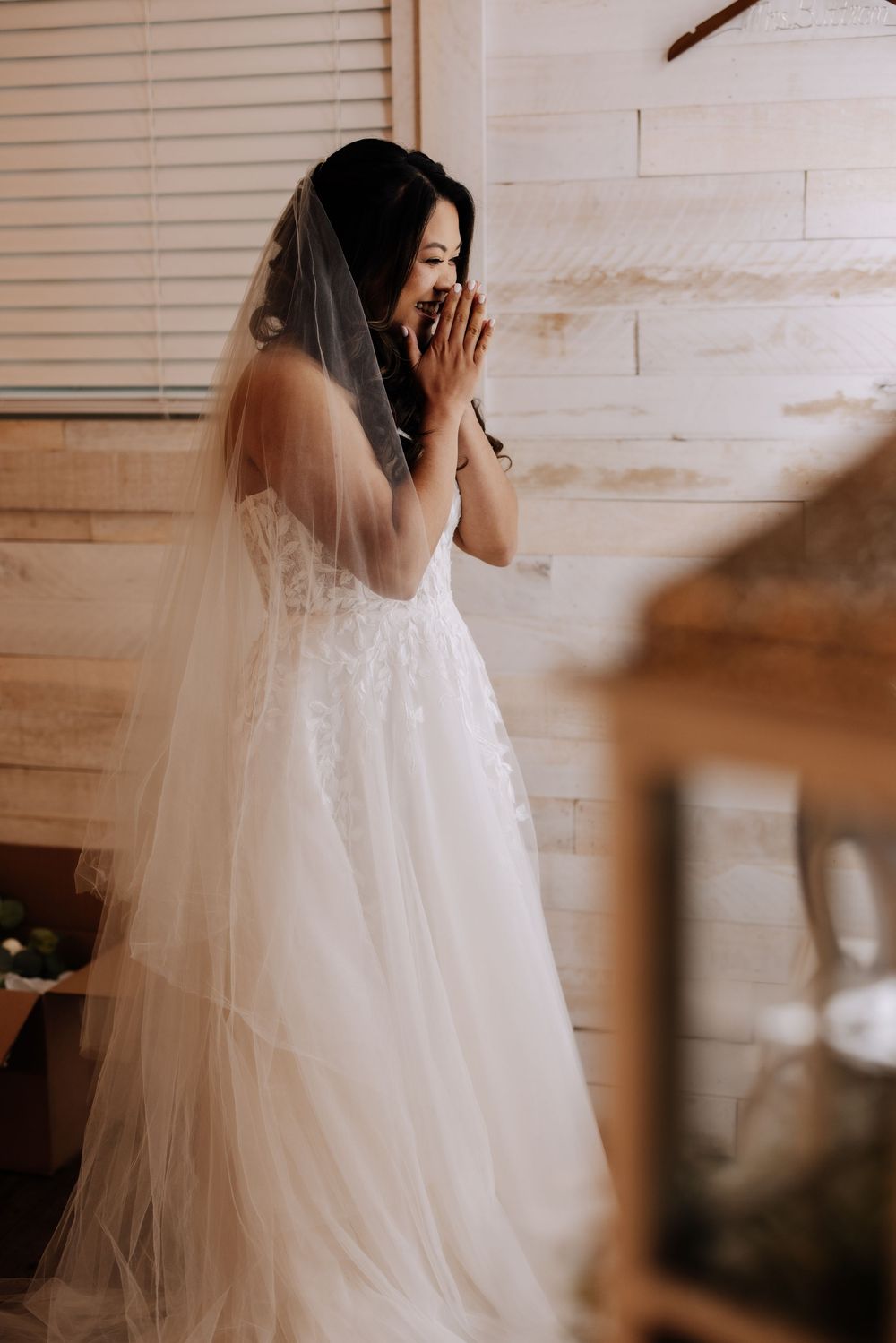 Bride in a white lace wedding gown and veil laughing joyfully in a rustic wooden room.