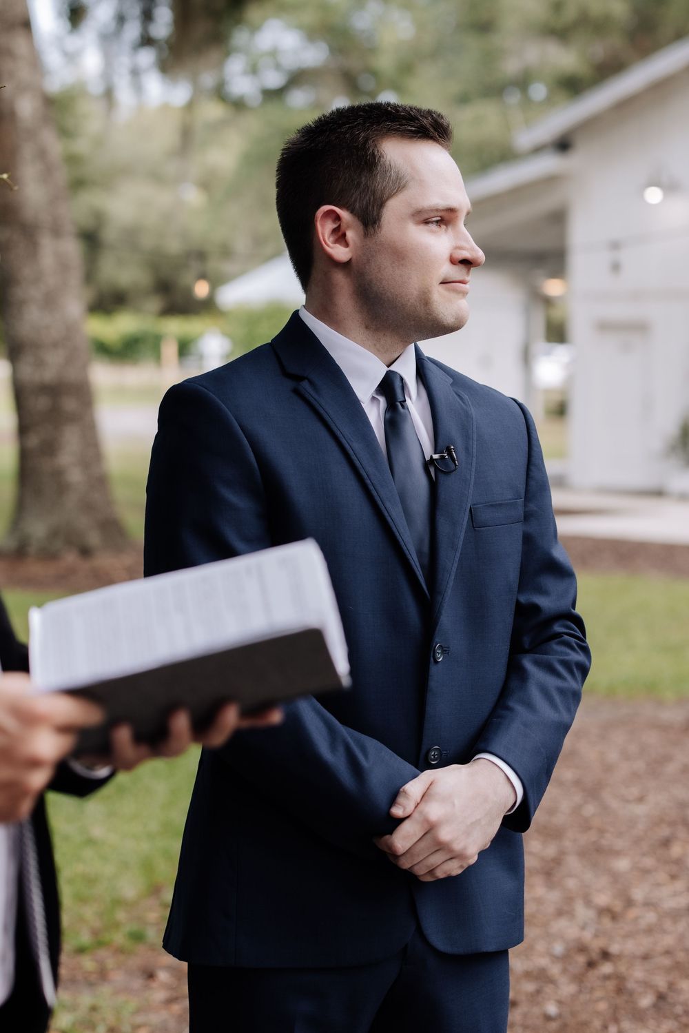 Groom in navy blue suit smiling during outdoor wedding ceremony, with officiant holding book in foreground.