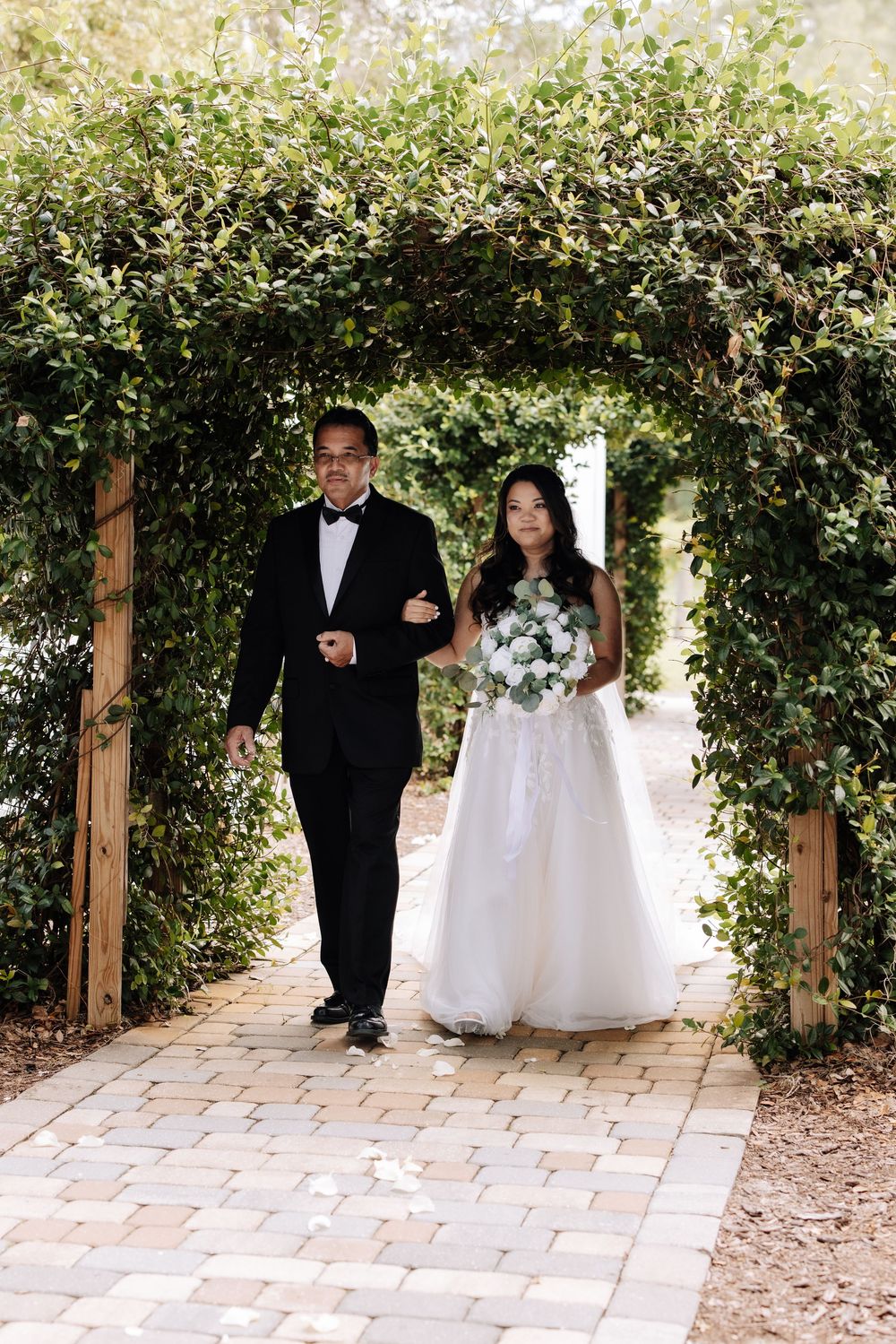 Father walking bride down a brick path under a lush green garden archway with bouquet.