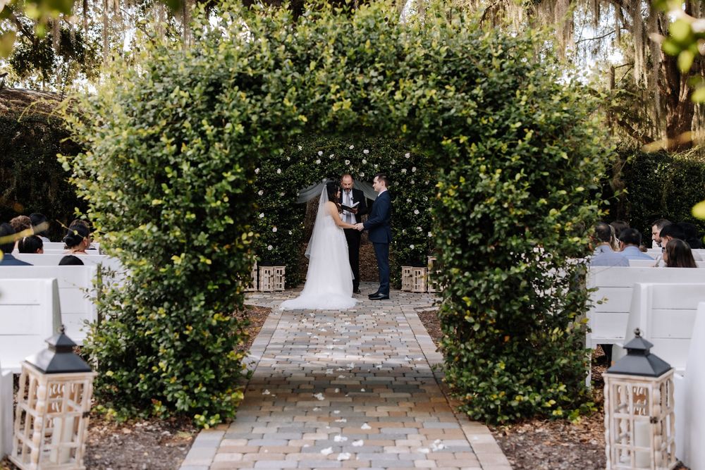 Couple exchanges vows under lush green arch at outdoor garden wedding ceremony with white pews lining brick aisle.