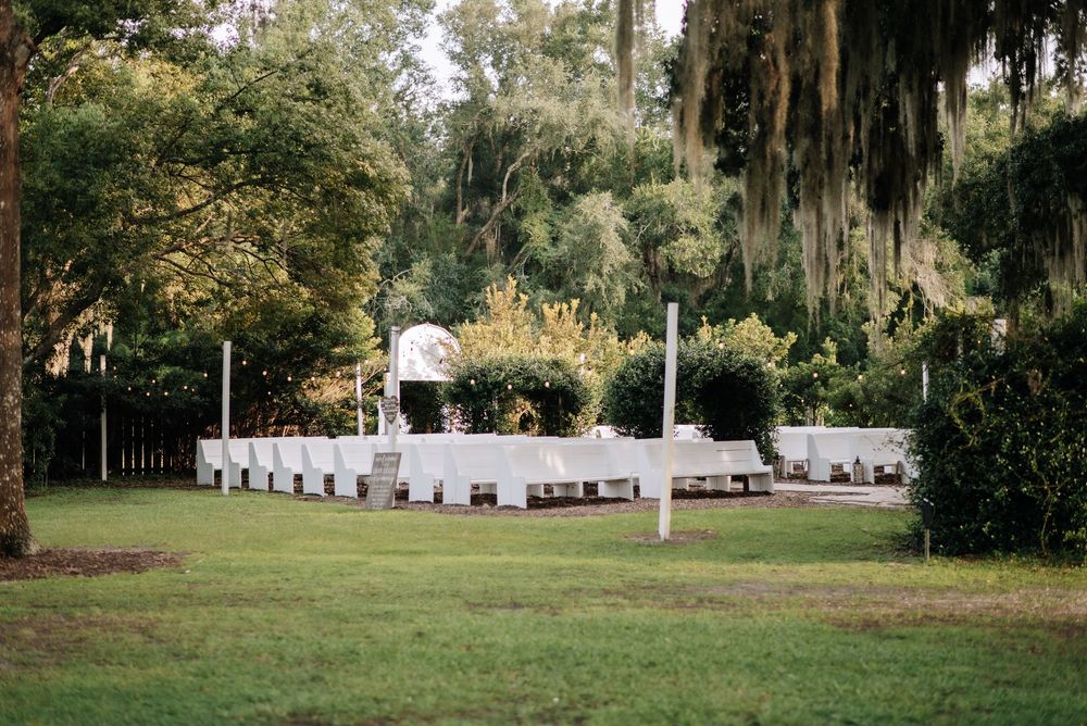 Outdoor wedding ceremony setup with white chairs and floral arch surrounded by lush green trees and Spanish moss.
