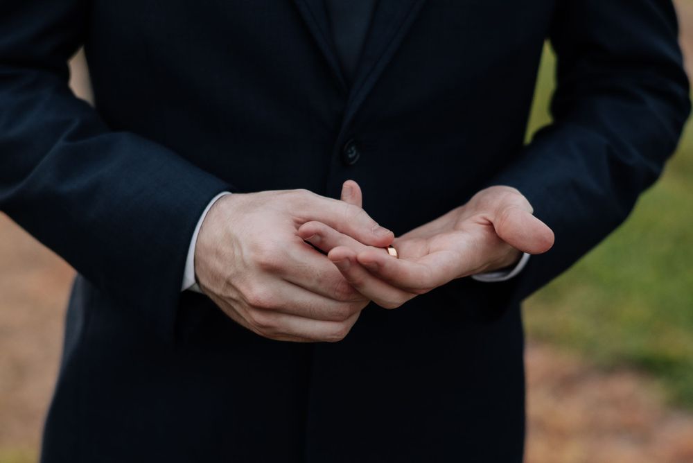 Groom in black suit holding wedding ring between fingers before ceremony.