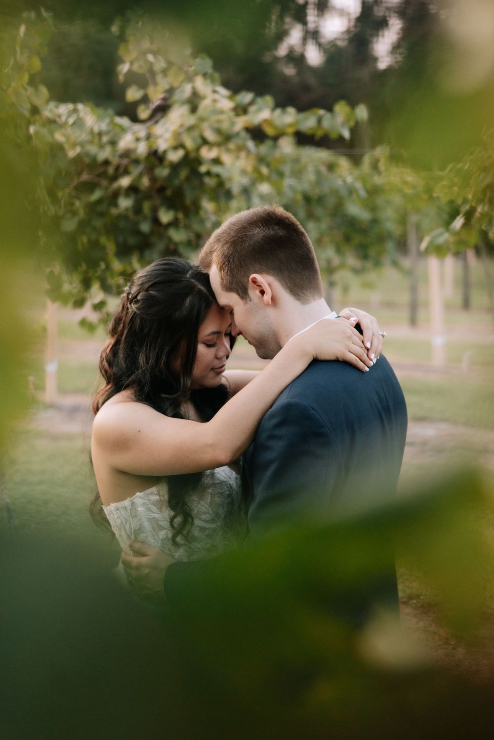 Newlyweds sharing an intimate forehead touch embrace surrounded by soft green foliage at outdoor wedding.