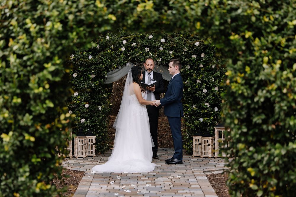 Bride and groom exchange vows under a floral arch surrounded by lush greenery at an outdoor wedding ceremony.