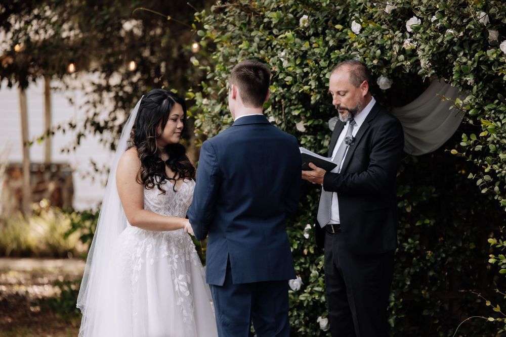 Bride and groom exchange vows outdoors with officiant, surrounded by lush greenery and white flowers.