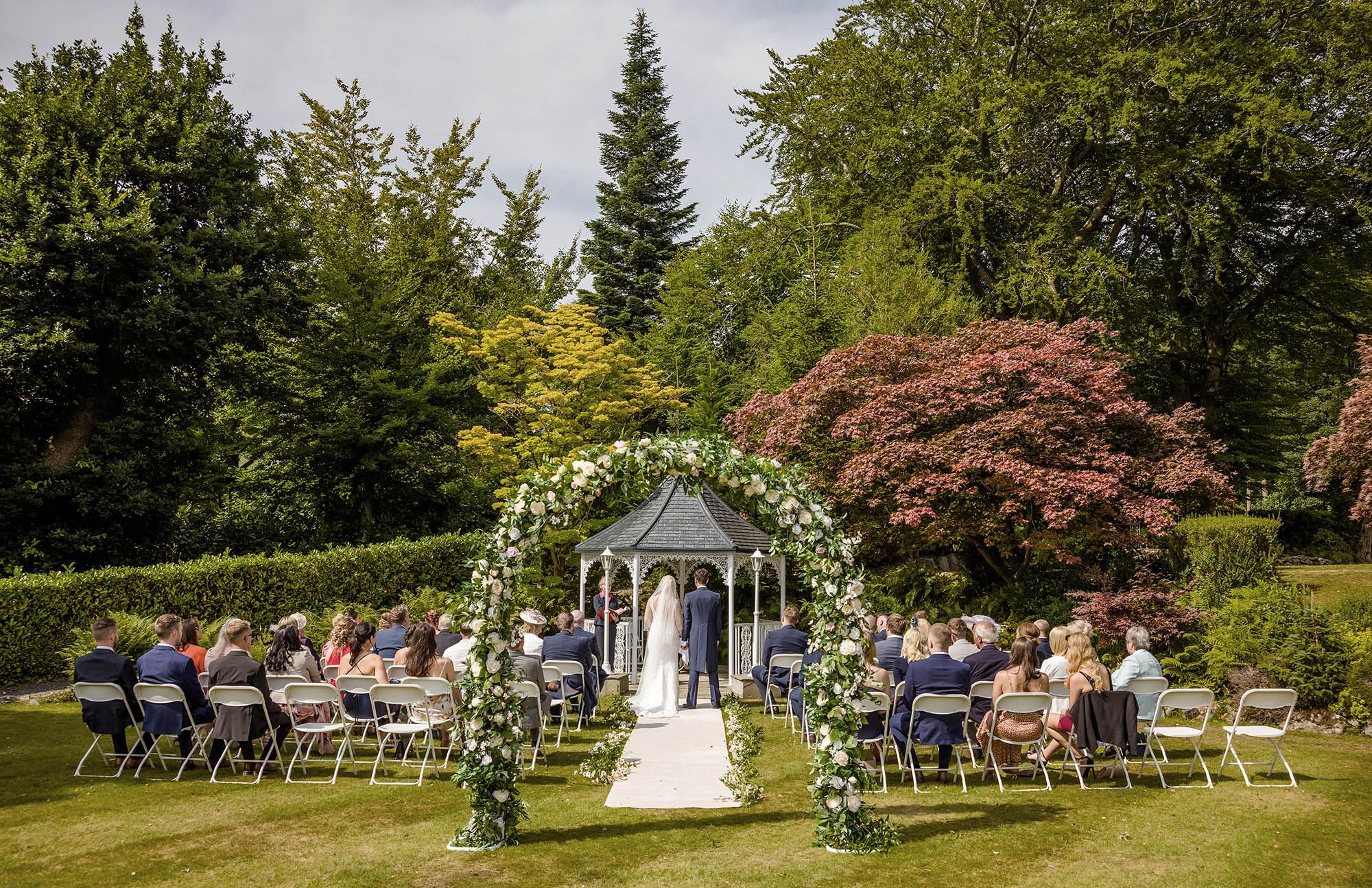 Outdoor wedding ceremony in a lush garden with floral arch, gazebo, and guests seated on chairs surrounded by trees.