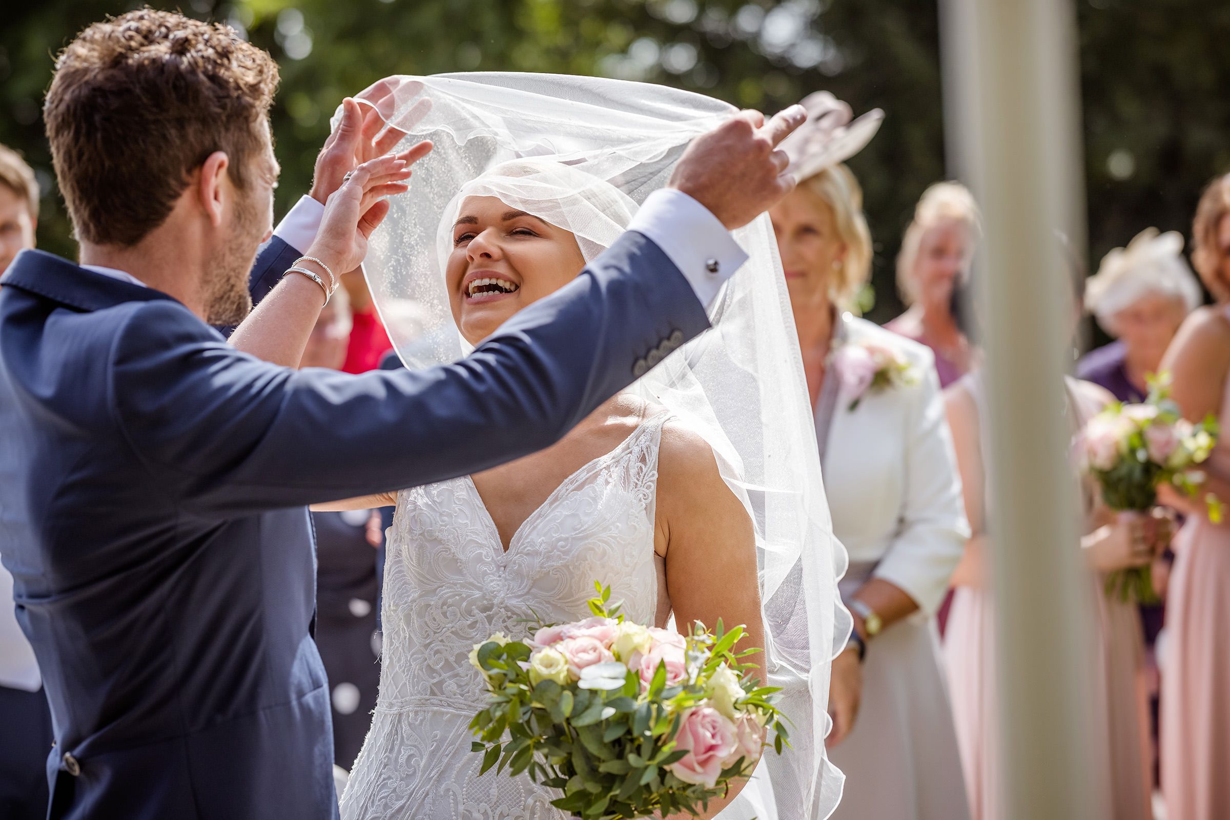 Groom lifting bride's veil outdoors at wedding ceremony while she laughs joyfully, guests watching in background.