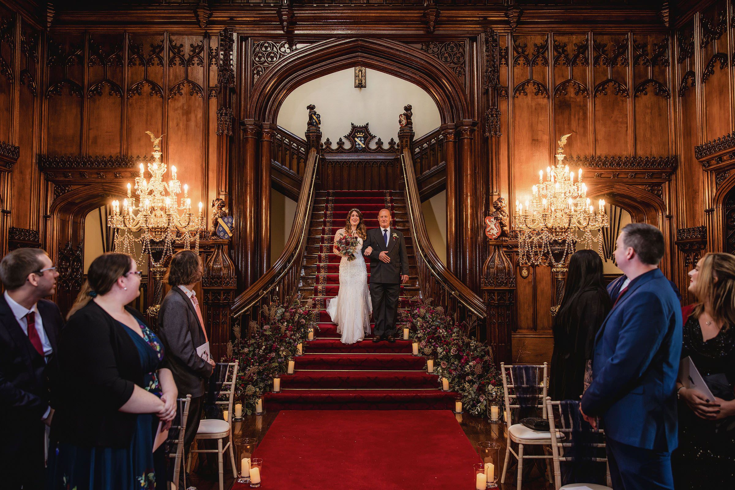 Bride and groom descend grand staircase with red carpet at elegant wood-paneled wedding ceremony venue with chandeliers.