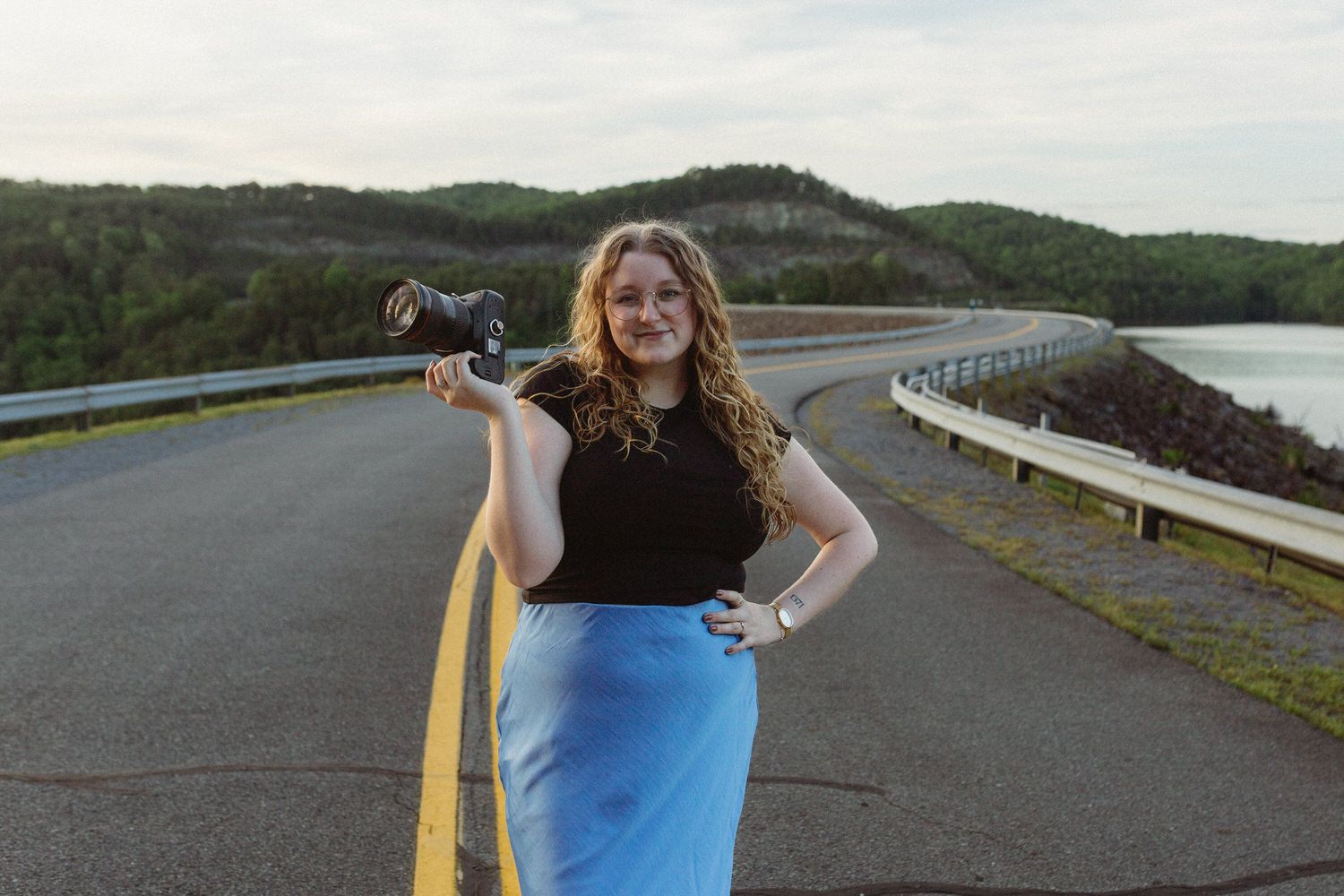 Elopement photographer stands on curved lakeside road holding camera wearing black top and bright blue skirt in the North Georgia Mountains