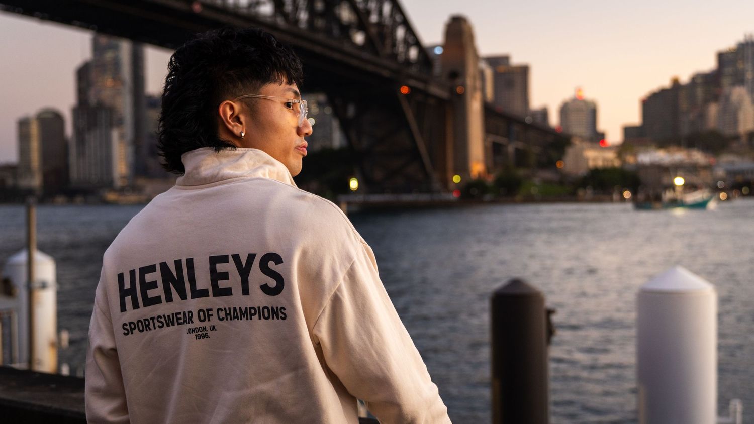 Person wearing white Henleys Sportswear jacket standing by Sydney Harbour Bridge at sunset.