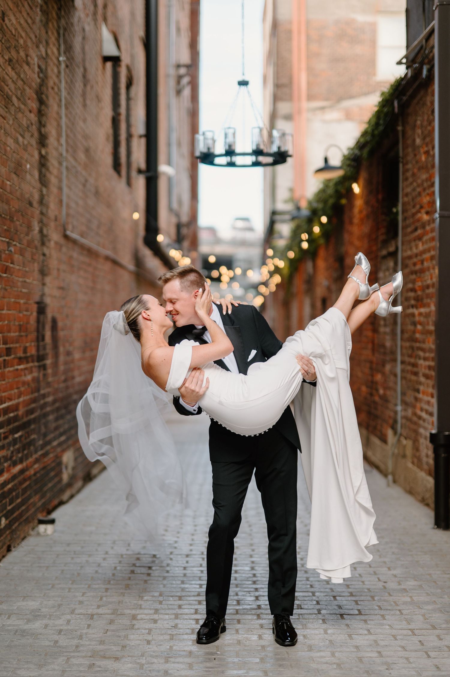 A bride and groom share a romantic moment, smiling at each other, she in white with floral hair accessories, he in a red suit.