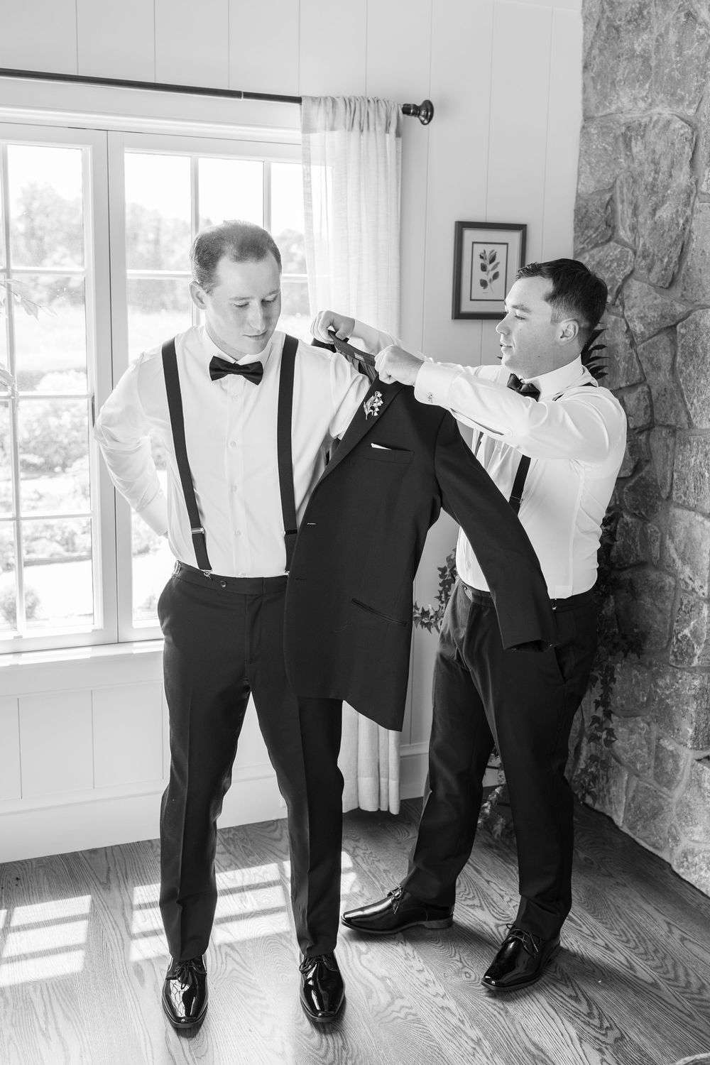 Black and white photo of groom putting on tuxedo jacket with help from groomsman before wedding ceremony.