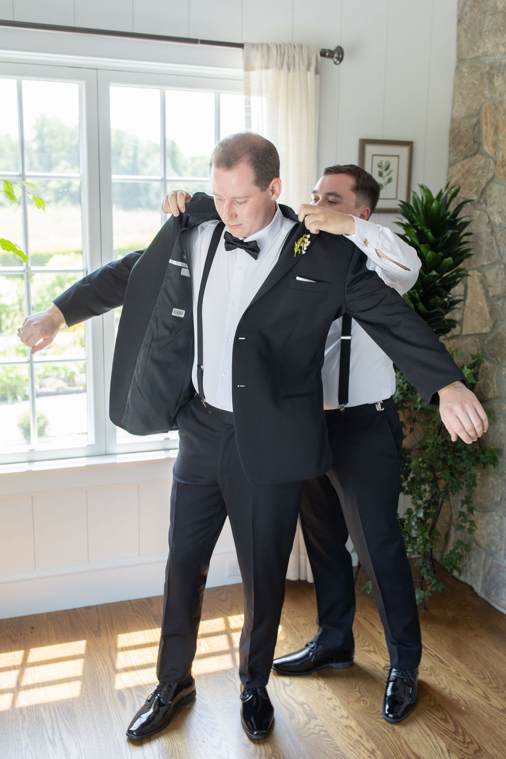 Groom being helped into black tuxedo jacket by groomsman before wedding ceremony indoors.