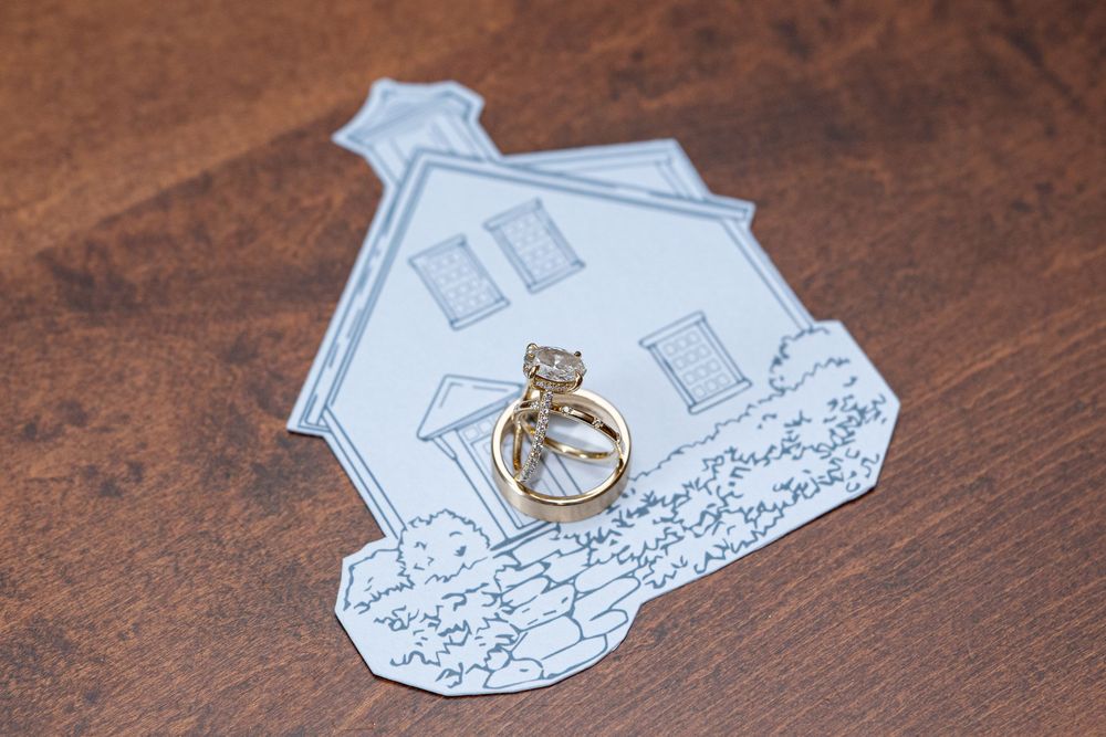 Gold diamond engagement ring and wedding band resting on a house-shaped card on a wooden surface.