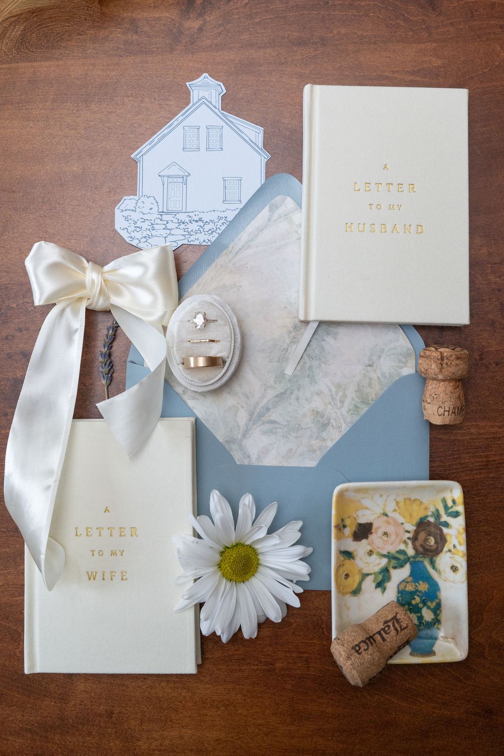 Wedding flat lay with vow books, ring box, daisy, blue envelope, and floral trinket dish on wood.