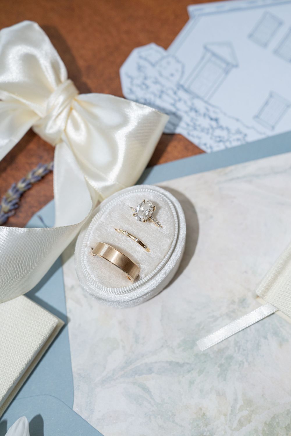 Gold wedding bands and diamond engagement ring in velvet box with white bow and blue wedding stationery.