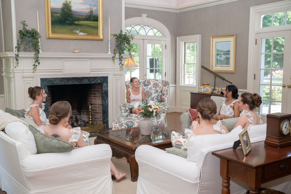 Bridesmaids in white dresses gather in an elegant living room with a fireplace during a bridal party getting ready session.