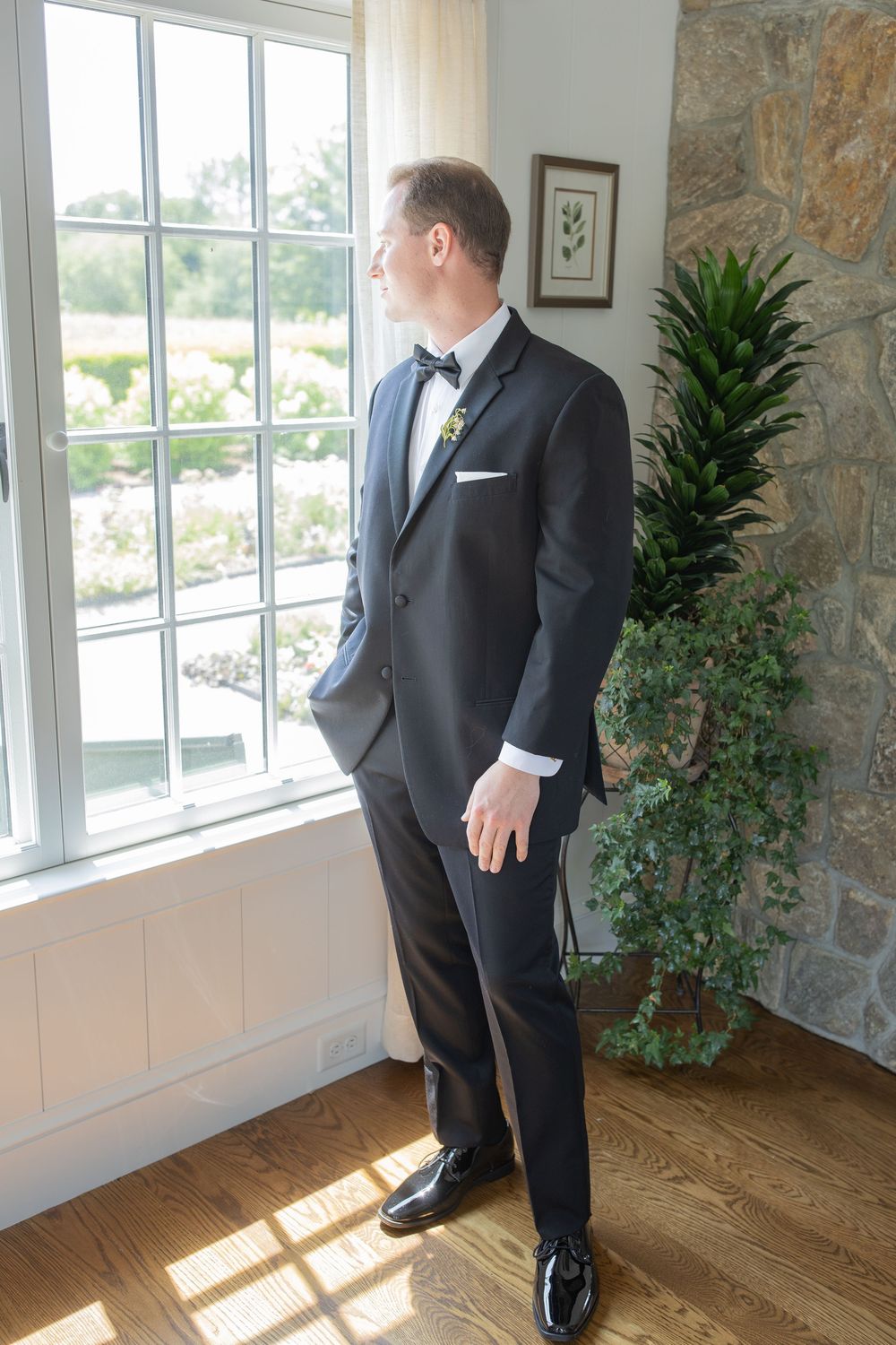 Groom in black tuxedo with bow tie stands by sunlit window in elegant room with stone wall and tropical plant.