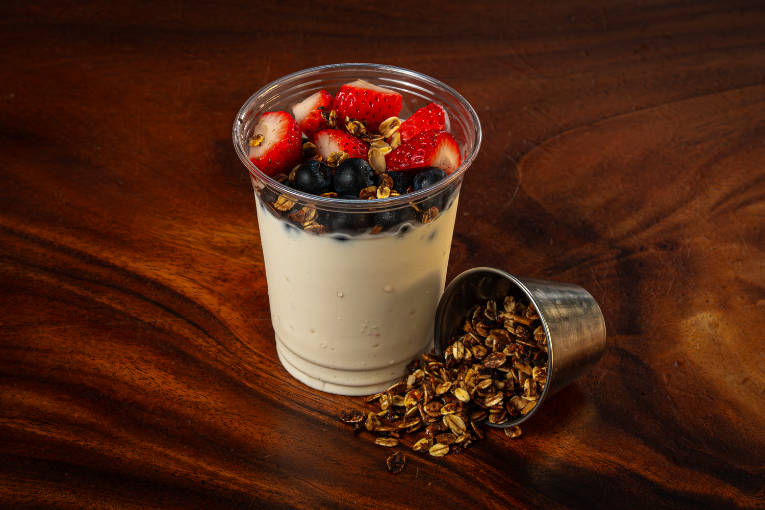 Creamy yogurt parfait topped with fresh strawberries and blueberries in a clear cup, with granola spilling from a metal cup.