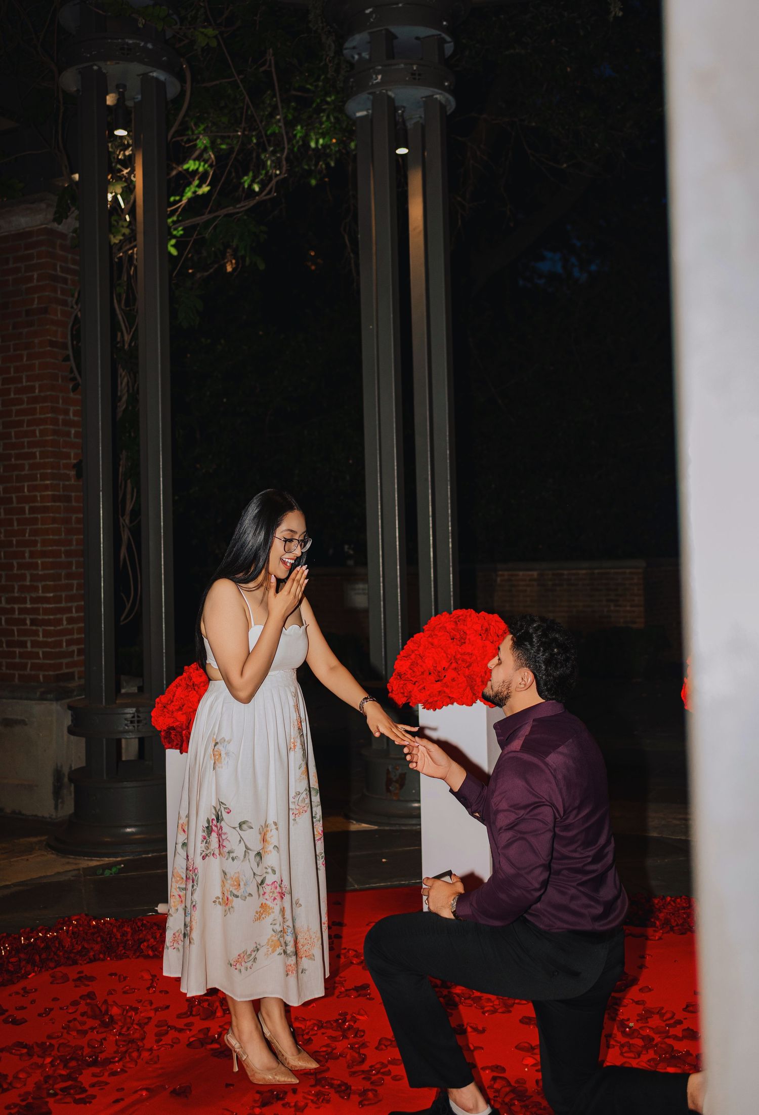 Man kneeling on red rose petals proposing to woman in floral dress at night with bouquet of red roses.