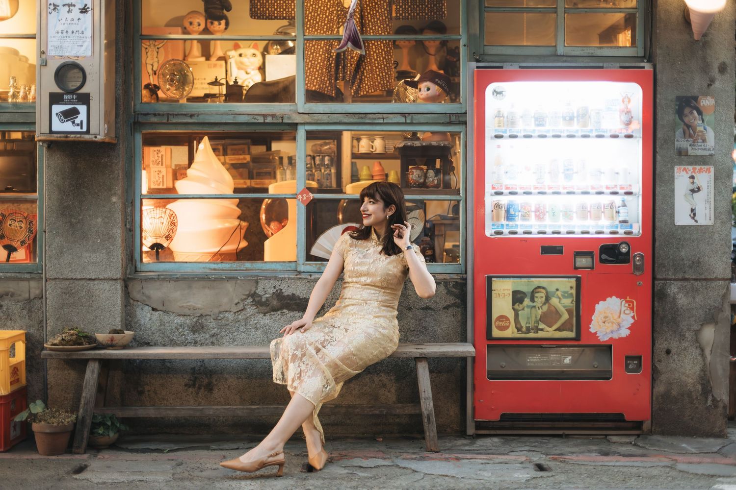 Woman in vintage dress sitting on bench beside red vending machine outside rustic shop.