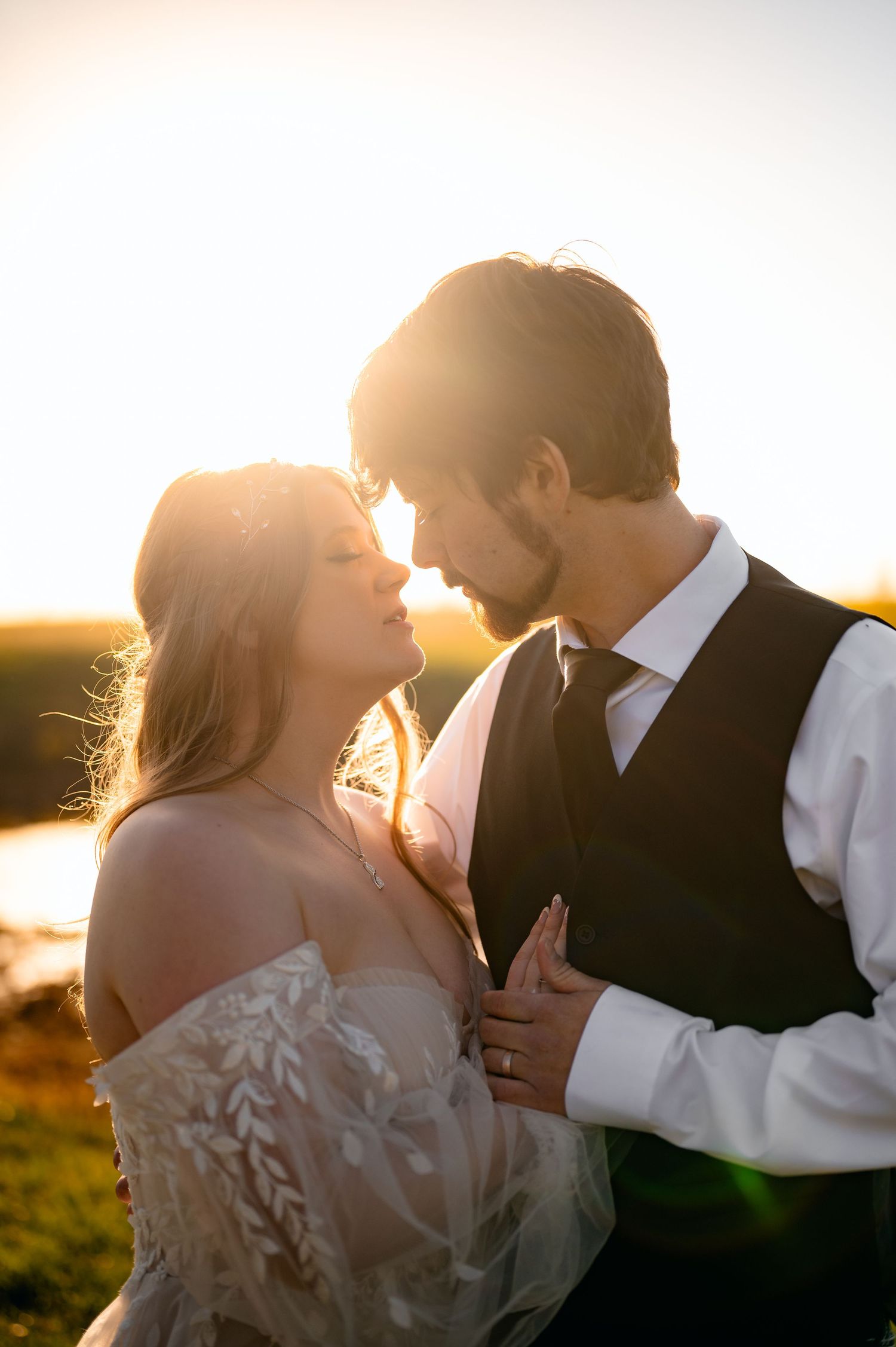 Interracial couple posing in a green field on their wedding day, bride in white gown holding bouquet, groom in black suit.