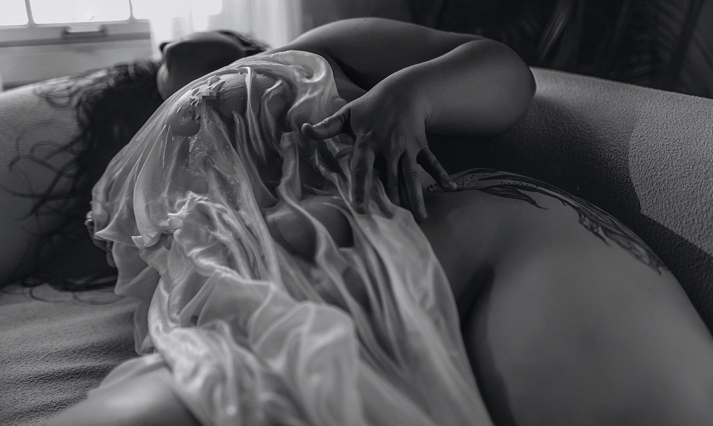 Black and white artistic photo of a woman draped in sheer fabric lying on a soft surface.