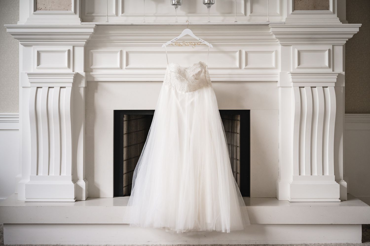 Elegant white wedding dress hanging on a hanger above a classic white fireplace mantel.