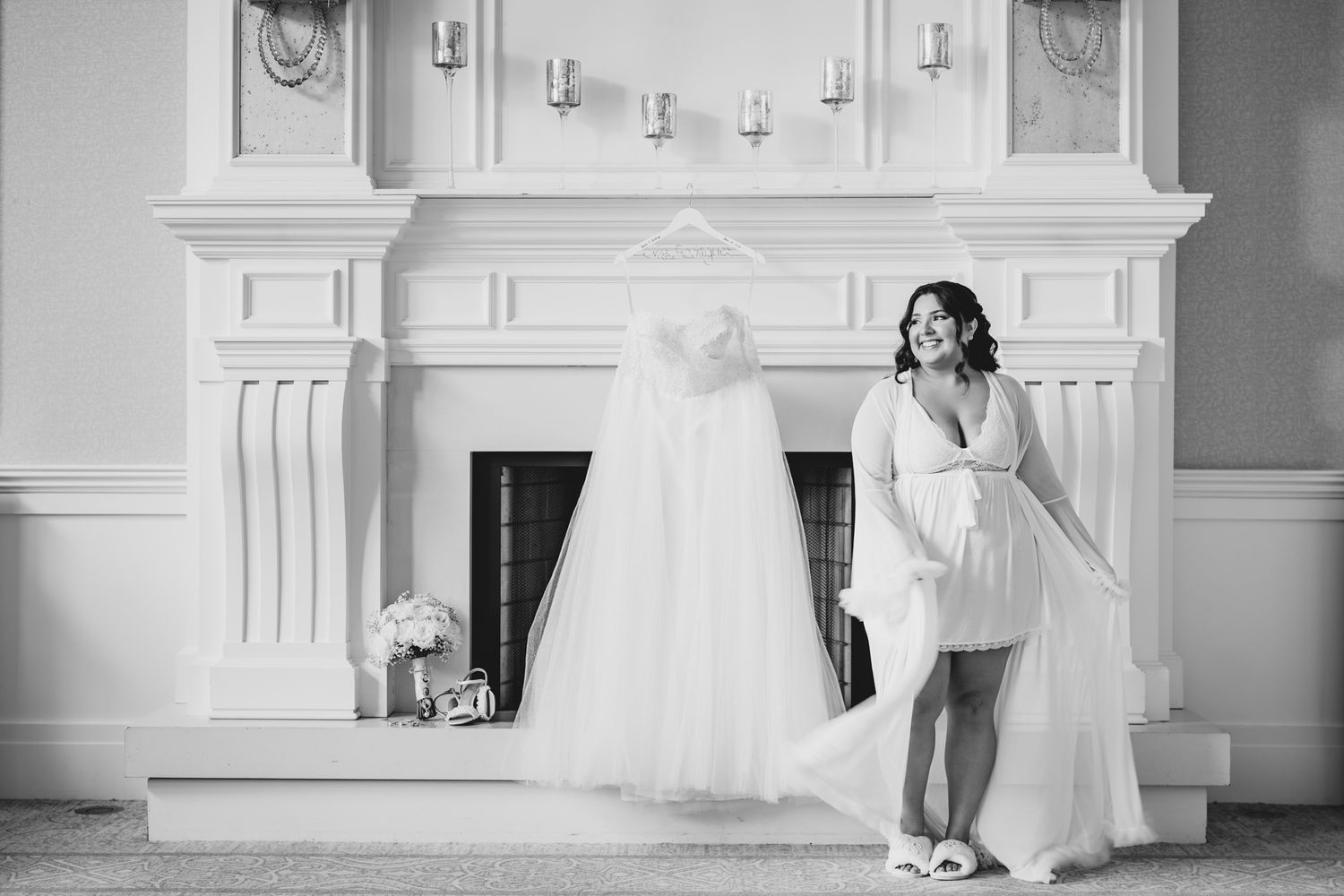 Bride in robe admiring her wedding gown hanging on fireplace mantel in elegant black and white bridal preparation photo.