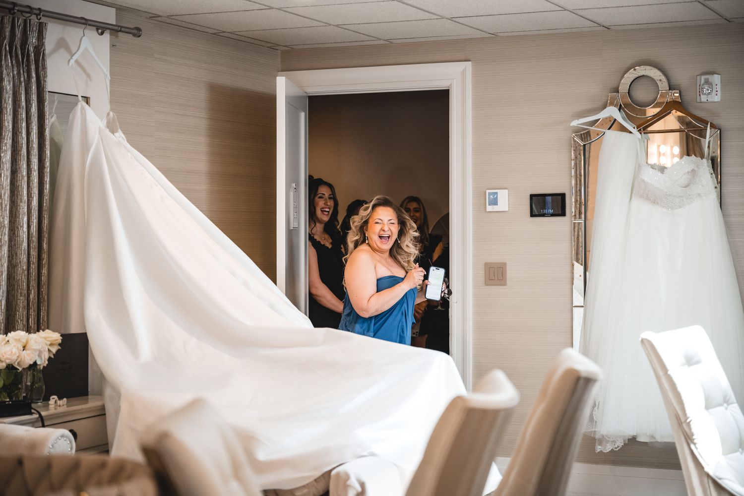 Bridesmaid in blue dress reacts with joy upon seeing the wedding gown displayed in a bridal suite.