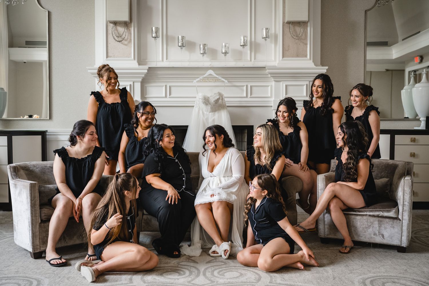 Bride in white robe poses with bridesmaids in black outfits near a wedding dress display in an elegant room.