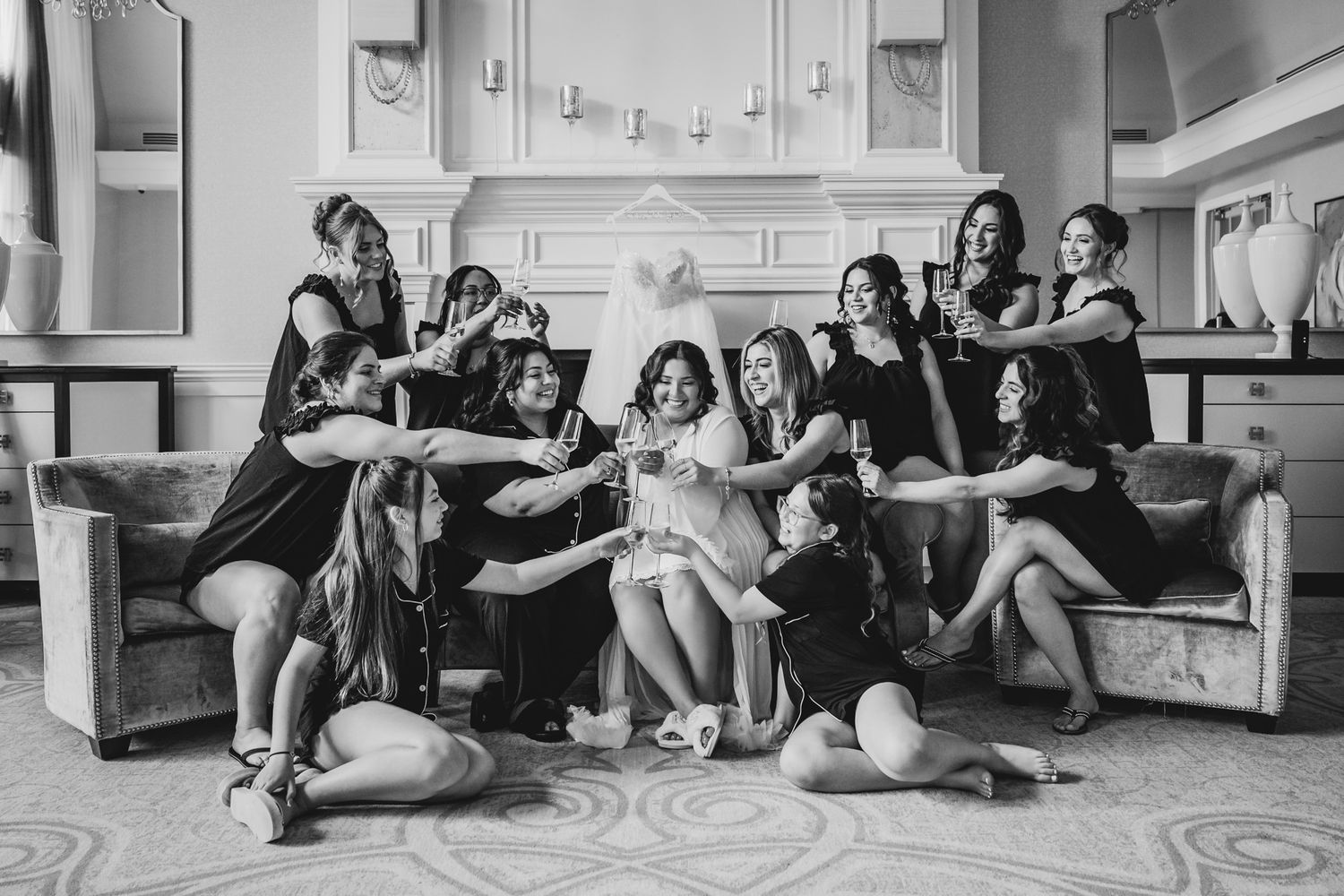 Black and white photo of bride and bridesmaids in black dresses celebrating together on a sofa in elegant venue.