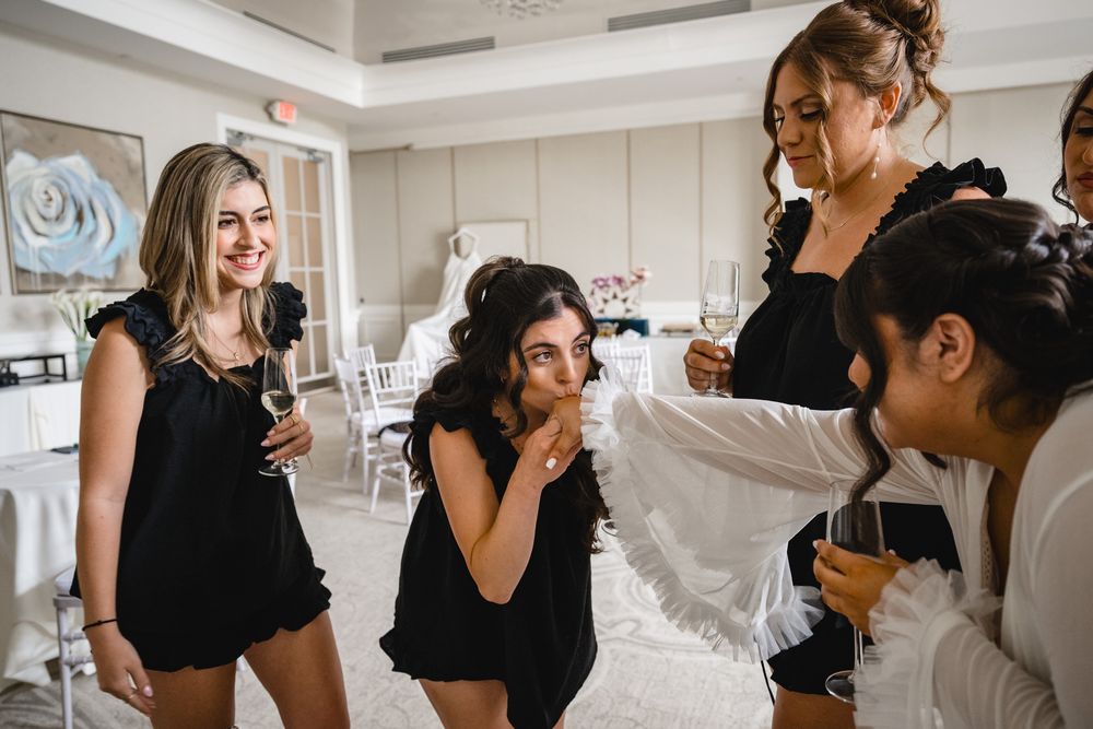 Bridesmaids in black outfits laughing and holding champagne glasses during bridal preparations in elegant white room.