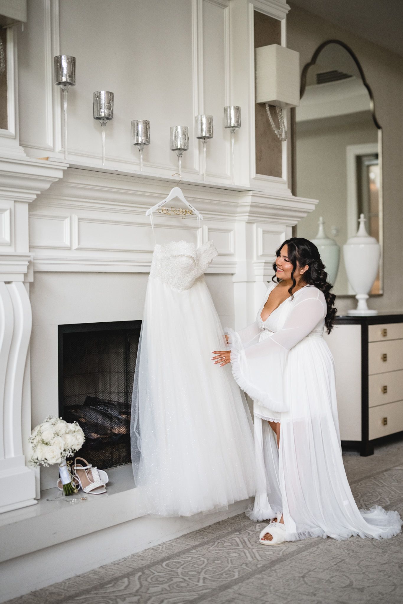 Bride in white robe admiring her wedding gown hanging over a fireplace in an elegant room.