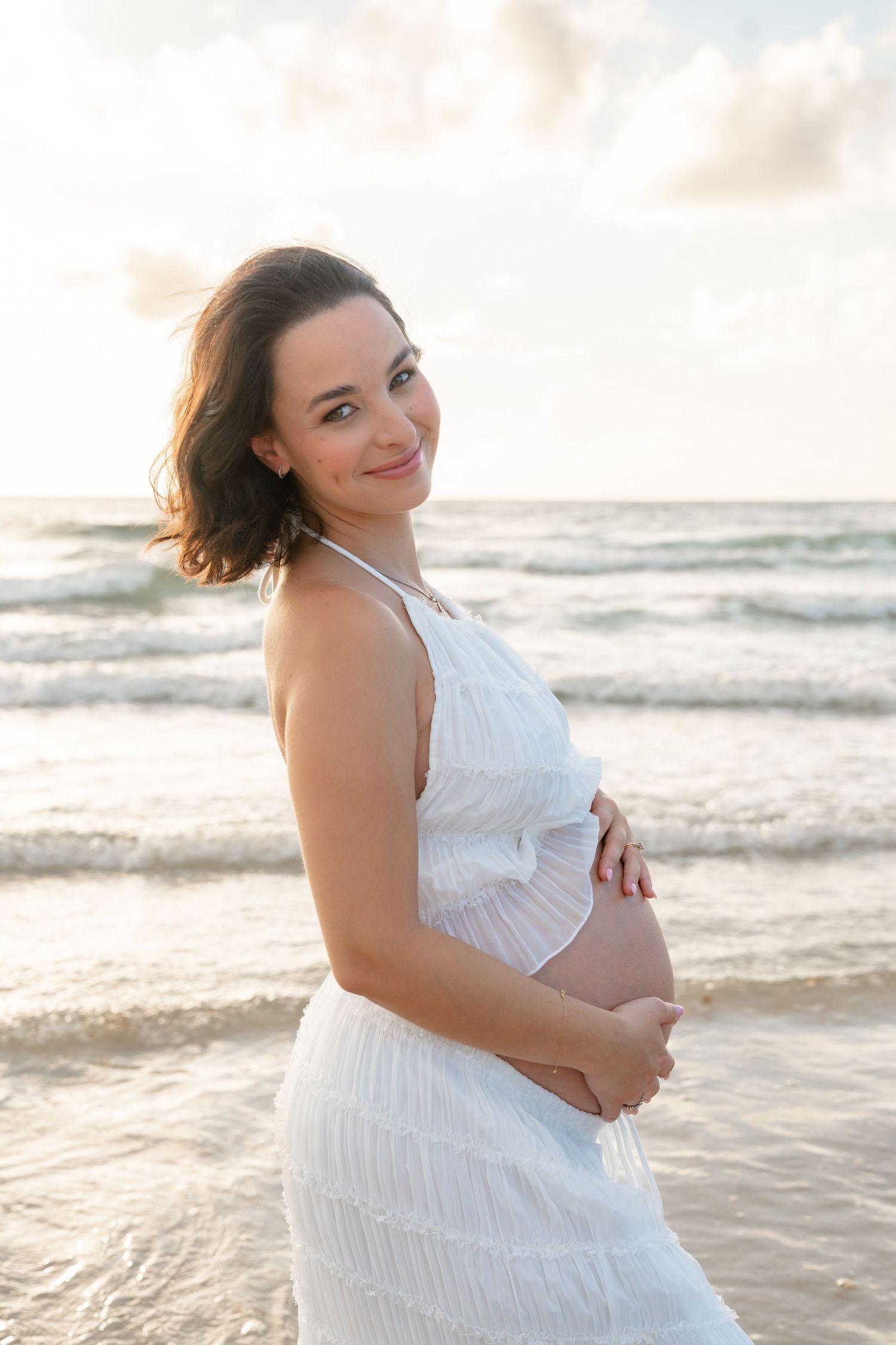 Smiling pregnant woman in white dress cradling baby bump on beach at sunset.