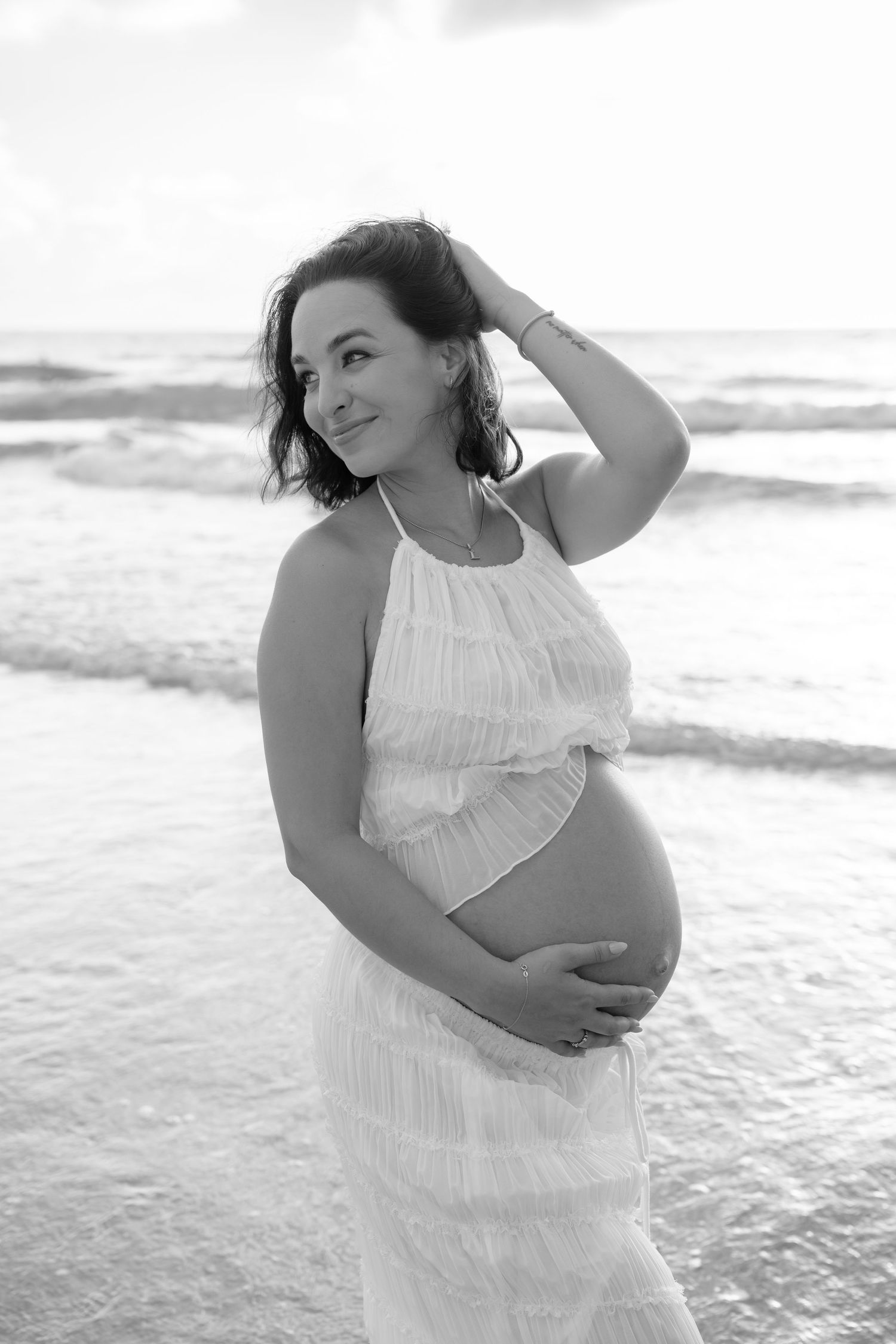 Black and white photo of a pregnant woman in a white dress cradling her belly on a beach with ocean waves behind her.
