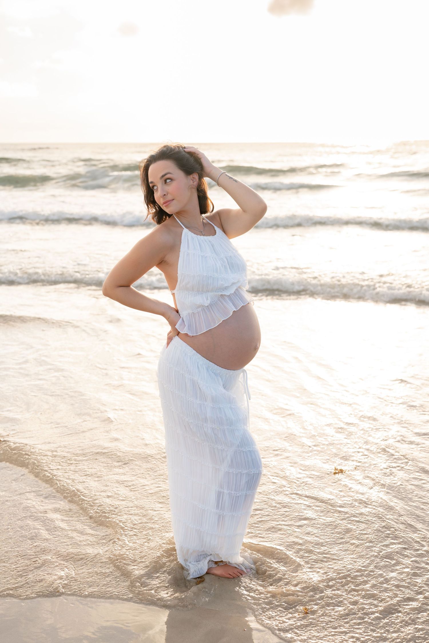 Pregnant woman in white dress standing on beach at sunset, cradling baby bump with waves in background.
