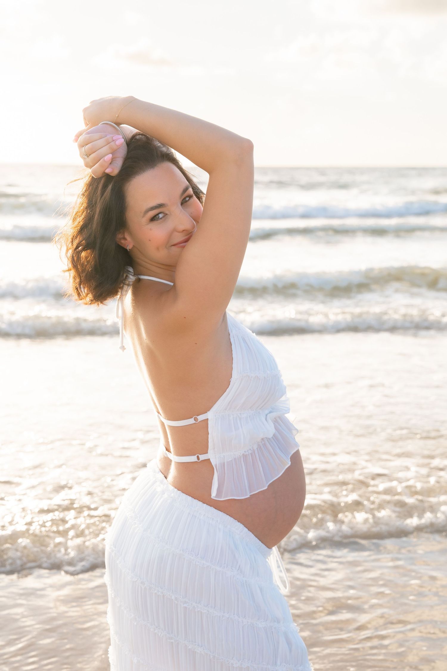 Pregnant woman in white backless dress posing on beach at sunset, smiling over shoulder with waves behind her.