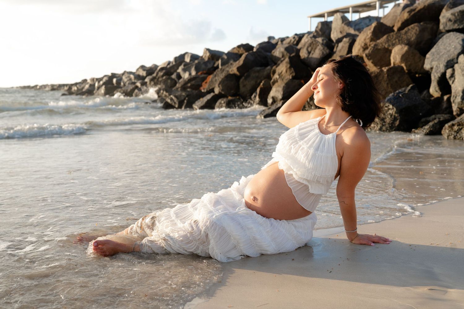 Pregnant woman in white dress sitting on sandy beach shore with rocky jetty and ocean waves in background.