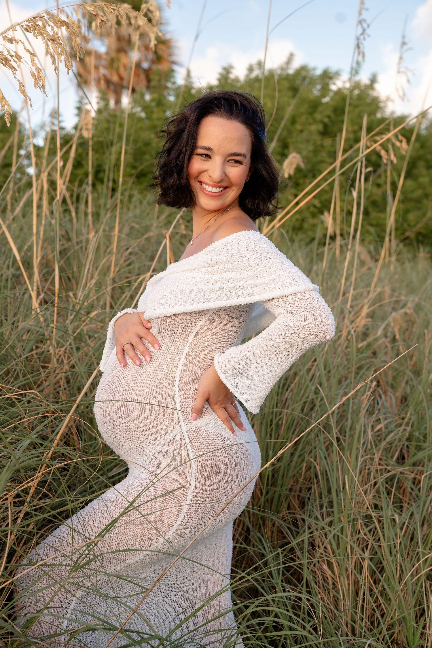 Smiling pregnant woman in white lace off-shoulder gown poses outdoors among tall grass at golden hour.