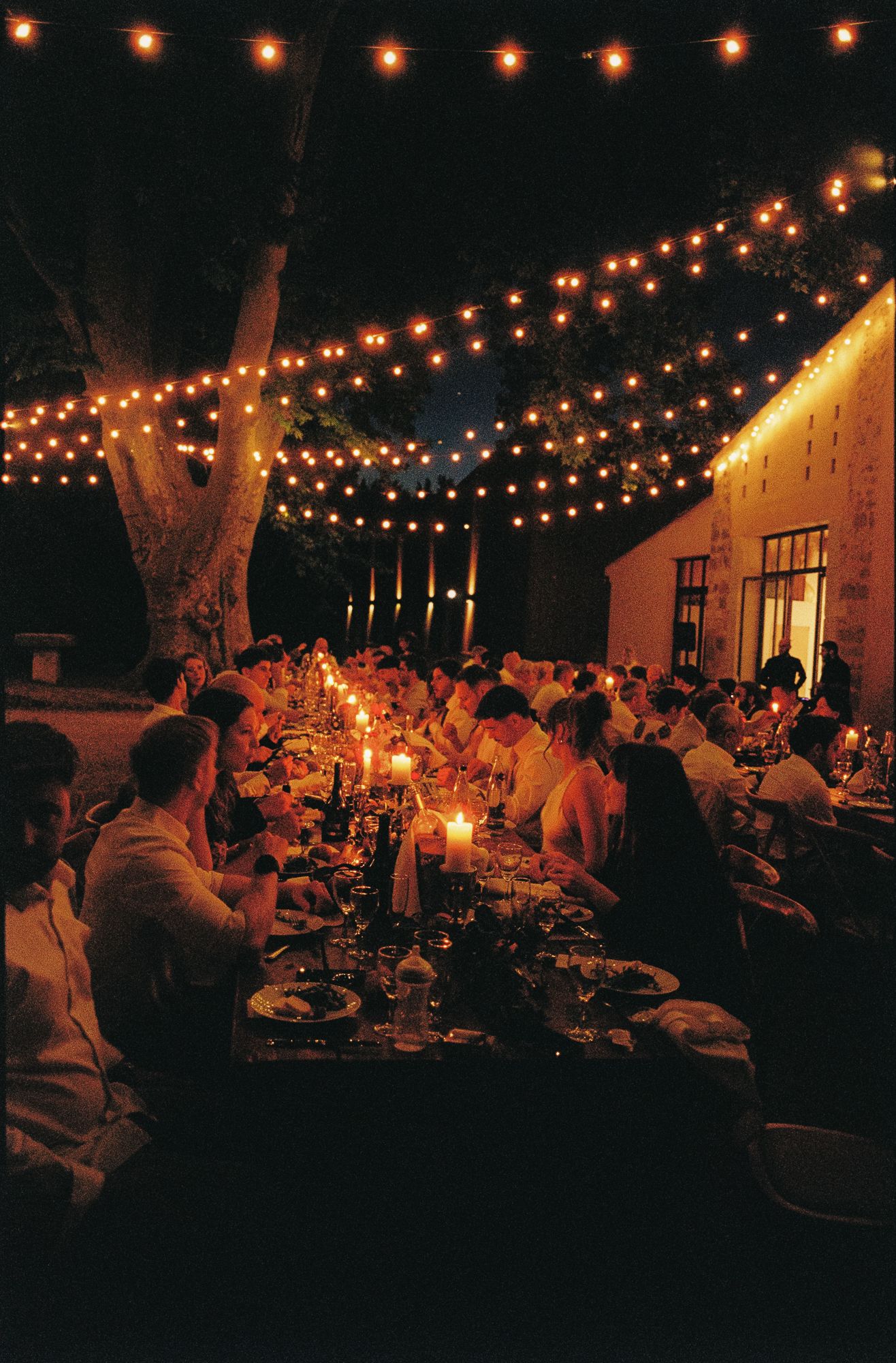 Outdoor evening dinner party with string lights and candles illuminating long tables of guests near a rustic building.
