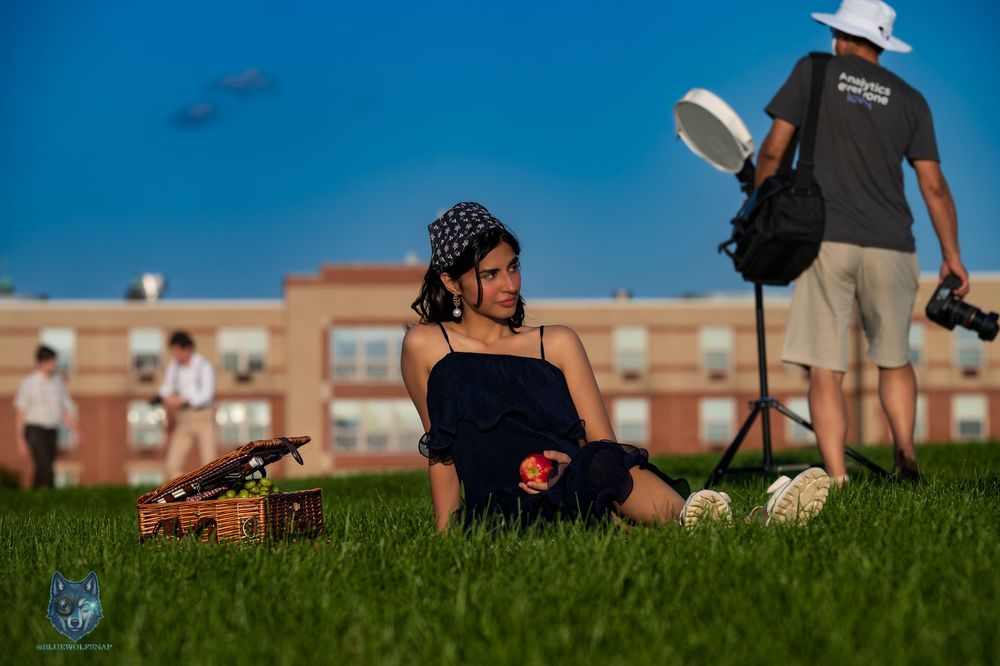 Woman in navy dress and bandana sitting on grass during an outdoor photoshoot with photographer in background. Chi.Shooters itsaqsaforyou