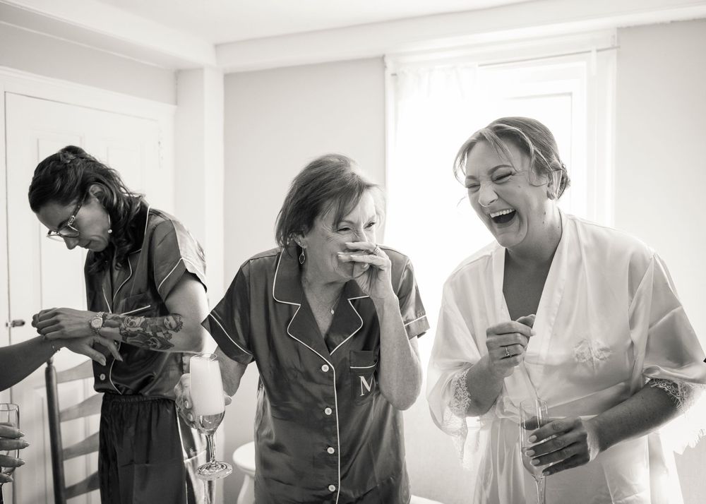 Three women in pajamas and robes laugh joyfully together while getting ready for a wedding in a bright room, in black and white.