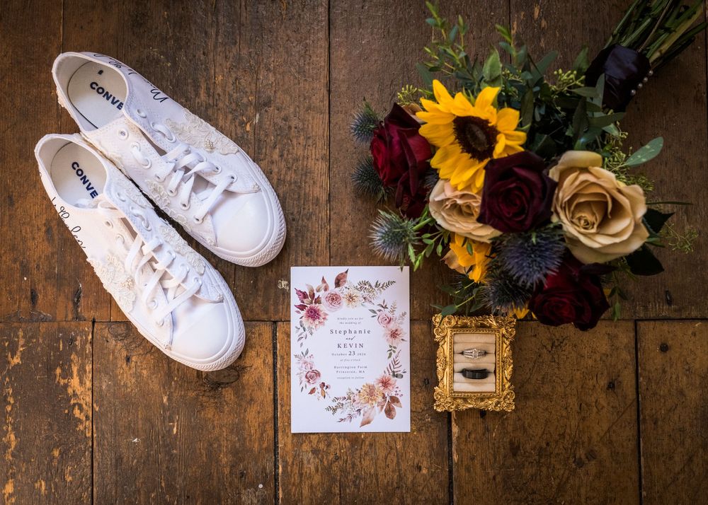 White Converse sneakers, rustic wedding invitation, and sunflower and burgundy rose bouquet arranged on a weathered wood floor.