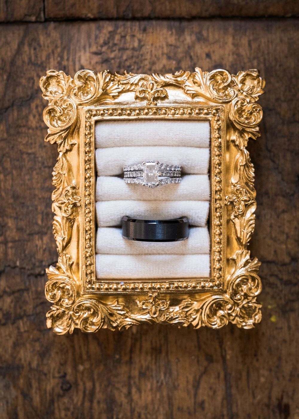 Diamond engagement ring and black wedding band displayed on cream cushions inside an ornate gold vintage picture frame on rustic wood.