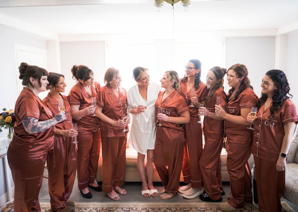 Bridesmaids in matching rust satin robes raise champagne glasses with the bride in a white robe in a bright getting-ready room.