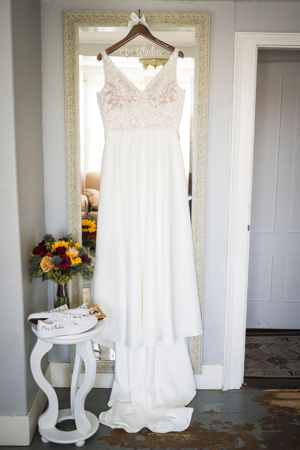 White lace-bodice wedding gown with long train hanging on a personalized hanger before an ornate mirror beside autumn bouquet.